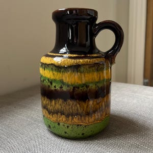 May include: A vintage ceramic pitcher with a dark brown top and handle, transitioning to bands of yellow, green, and brown, and ending with a green base. The pitcher has a glossy finish and a rounded body.