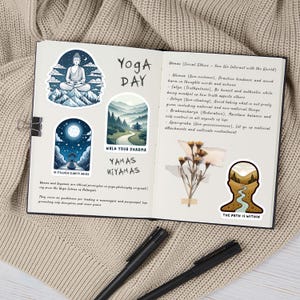 May include: An open journal featuring stickers of a meditating Buddha, a mountain path, a night scene with a full moon, and a landscape silhouette. The pages contain handwritten text about Yoga Day, Yamas, and Niyamas, alongside a dried flower.