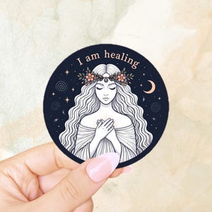 Puede incluir: Una pegatina redonda con un fondo azul oscuro que presenta a una mujer con largo cabello blanco ondulado, una corona de flores y el texto "I am healing". La mujer tiene las manos juntas sobre el pecho, rodeada de estrellas y una luna creciente.