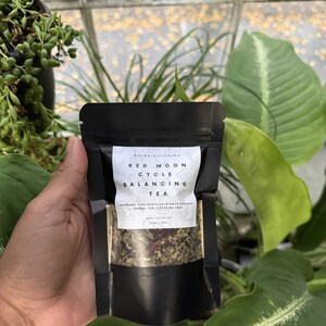 May include: A black pouch of herbal tea labeled "Red Moon Cycle Balancing Tea." The label includes the ingredients: raspberry leaf, hibiscus, ginger, and spearmint. The tea is a mix of green and red herbs.