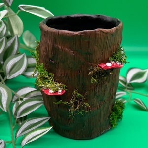 Könnte beinhalten: Ein brauner Keramiktopf in Form eines Baumstamms mit grünen Moos- und roten Pilz-Akzenten.