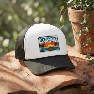 May include: A white and black trucker hat featuring a rectangular patch on the front panel. The patch displays the text OCEANSIDE CALIFORNIA above a graphic of a sunset, palm trees, and a pier over water. The hat has a black brim and mesh back.