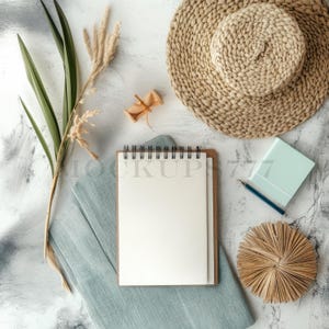 Puede incluir: Una maqueta de plano plano con un cuaderno en espiral en blanco con una cubierta marrón, una tela azul, un sombrero de paja tejido, una pequeña caja de madera, un bloc de notas azul, un lápiz y un círculo de paja tejido.