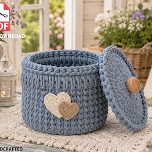 May include: A round, blue crocheted basket with a matching lid featuring a light wooden knob. The exterior of the basket is decorated with two small, layered heart-shaped appliques in cream and beige tones, showcasing a textured yarn stitch.