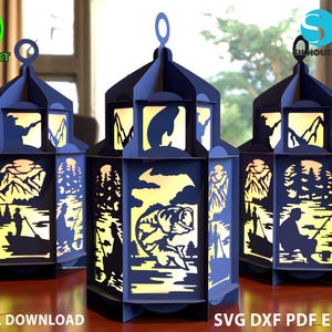 May include: Three blue paper lanterns with intricate cut-out designs depicting a fishing scene. The lanterns are illuminated from within, casting a warm glow. The designs include a fisherman in a boat, a bass fish, and a mountain landscape.