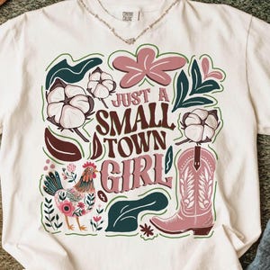 Pu&ograve; includere: T-shirt bianco con un design grafico colorato. Il design include il testo "Just a Small Town Girl" con illustrazioni di cotone, uno stivale da cowboy rosa, un gallo ed elementi floreali. La maglietta ha uno scollo tondo.