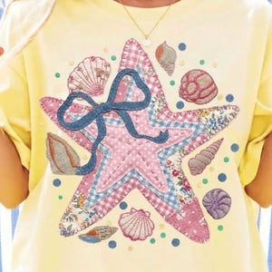 May include: Yellow t-shirt featuring a large, embroidered starfish design. The starfish is pink with a blue bow and surrounded by seashell illustrations. The shirt has a relaxed fit and a crew neck.