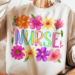 以下が含まれることがあります： クリーム色のスウェットシャツ。カラフルな縁取り文字で「NURSE」の文字が描かれ、オレンジ、ピンク、黄色、紫色の水彩画の花で囲まれています。デザインは衣服の前面中央に配置されています。