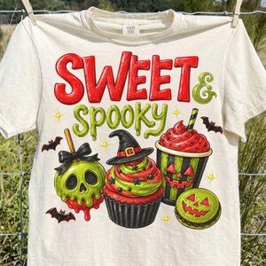 May include: A cream-colored t-shirt featuring the red and green text SWEET & SPOOKY. The graphic includes a green skull candy apple, a cupcake topped with a witch hat, a jack-o'-lantern drink cup, a green macaron, and small black bats.