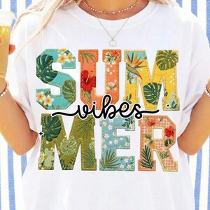 Könnte beinhalten: Weißes T-Shirt mit dem Schriftzug "SUMMER vibes" in bunten, floral gemusterten Buchstaben. Die Buchstaben sind mit einem Stichmuster umrandet. Das Shirt hat eine lockere Passform und ist für warmes Wetter geeignet.
