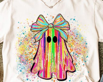 May include: White t-shirt featuring a colorful ghost design with a large bow. The ghost has vertical stripes in various colors, including pink, yellow, and green. The design is accented with gold glitter and colorful paint splatters.