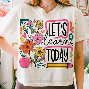 May include: Cream-colored t-shirt with a colorful floral design and the text "LET'S learn TODAY". The graphic includes pink, orange, and yellow flowers, a pink apple, and a yellow pencil.