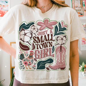 Pu&ograve; includere: T-shirt color crema con un design grafico. Il design presenta la scritta "JUST A SMALL TOWN GIRL", uno stivale da cowboy rosa, un gallo, batuffoli di cotone ed elementi floreali in rosa, verde e marrone.