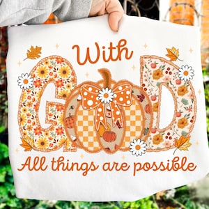 May include: White sweatshirt with the text "With God All things are possible." The design features a pumpkin with a daisy bow, surrounded by the letters "G" and "D" filled with floral patterns. Autumn leaves and daisies complete the design.