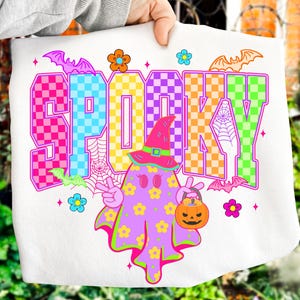 May include: White Halloween-themed tote bag with the word "SPOOKY" in colorful checkered letters. A cartoon ghost with a witch hat and a jack-o'-lantern bucket is featured. Bats and flowers decorate the design.