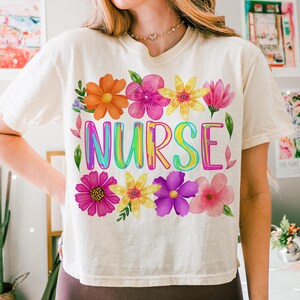 以下が含まれることがあります： クリーム色のTシャツ。レインボーカラーの「NURSE」の文字がプリントされ、オレンジ、ピンク、黄色、紫色の水彩画の花で囲まれています。 ゆったりとしたフィット感で、カジュアルなスタイルです。
