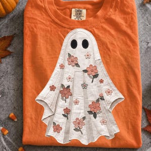Puede incluir: Una camiseta naranja con una aplicación central de fantasma hecha de tela color blanco roto con costuras verticales. El fantasma tiene dos ojos ovalados negros y está decorado con varias flores de tela pequeñas en rosa y melocotón y hojas verdes.