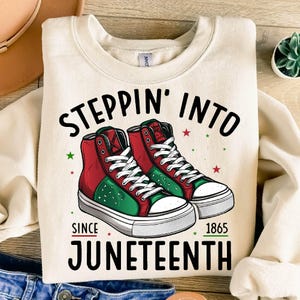 Może przedstawiać: Kremowa bluza z napisem "Steppin' Into Juneteenth Since 1865". Projekt przedstawia parę wysokich trampek w kolorze czerwonym, zielonym i czarnym. Trampki mają białe sznurowadła i podeszwy, z czerwonymi i zielonymi gwiazdami.