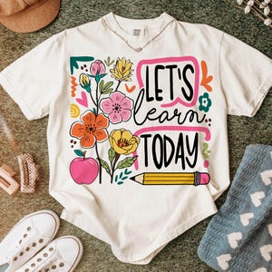 May include: Cream-colored t-shirt with a colorful floral design and the text "LET'S learn TODAY". The design includes an apple, pencil, and various flowers in shades of pink, orange, and yellow. The shirt is a casual, short-sleeved style.