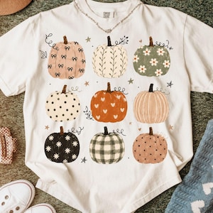 May include: A white short-sleeve t-shirt featuring a graphic print of nine pumpkins in three rows. The pumpkins display various patterns including bows, leaves, daisies, polka dots, hearts, stripes, and plaid in shades of orange, green, and black.