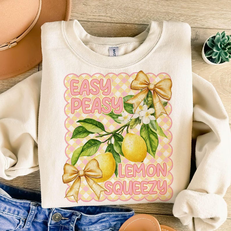 Pu&ograve; includere: Una felpa bianca con un design grafico rosa e giallo con limoni, un albero di limoni e il testo "Easy Peasy Lemon Squeezy".