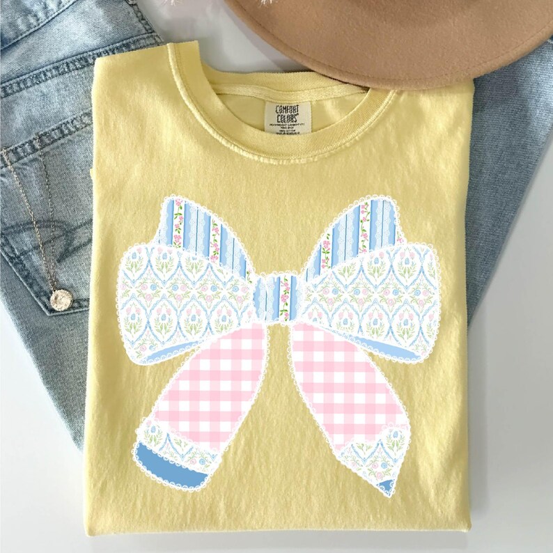 May include: A yellow t-shirt featuring a large patchwork bow design. The bow is made of blue, pink and white fabric with a floral pattern. The shirt is a comfortable fit and is ideal for everyday wear.