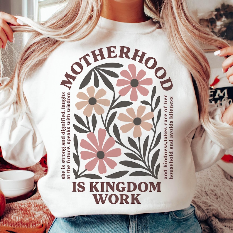 Puede incluir: Sudadera blanca con un dise&ntilde;o floral y el texto "MOTHERHOOD IS KINGDOM WORK" en letras marrones. El texto "she is strong and dignified, laughs at the future, speaks with wisdom and kindness, takes care of her household and avoids idleness" est&aacute; impreso en marr&oacute;n en el lado izquierdo del dise&ntilde;o.