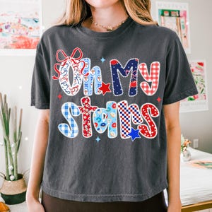 May include: Dark gray t-shirt with the phrase "Oh My Stars" in a colorful, patriotic design. The text features red, white, and blue patterns, including stars, gingham, and floral elements, perfect for celebrating the USA.