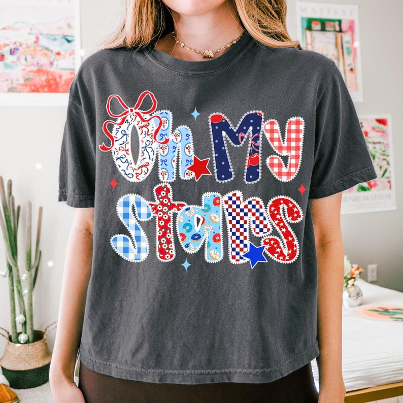 May include: Dark gray t-shirt with the phrase "Oh My Stars" in a colorful, patriotic design. The text features red, white, and blue patterns, including stars, gingham, and floral elements, perfect for celebrating the USA.