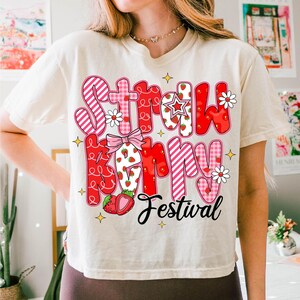 May include: A white t-shirt with a red and pink graphic that says "Strawberry Festival". The graphic is made up of different patterned letters and includes strawberries and flowers.
