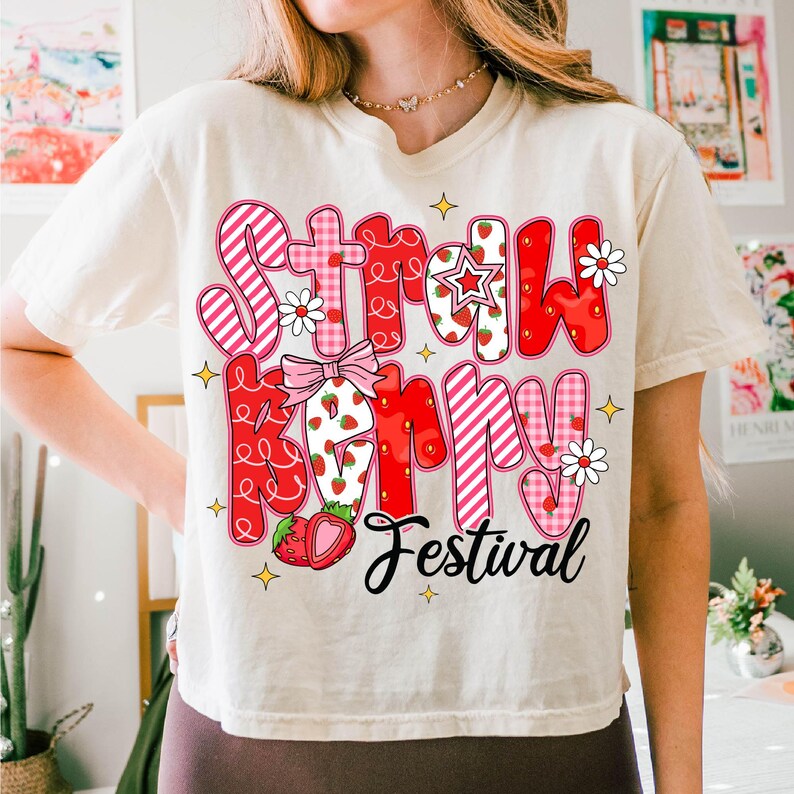 May include: A white t-shirt with a red and pink graphic that says "Strawberry Festival". The graphic is made up of different patterned letters and includes strawberries and flowers.