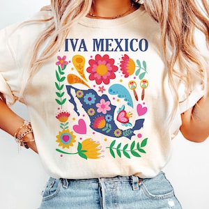 May include: A white t-shirt with a colorful graphic design featuring a stylized outline of Mexico with flowers, hearts, and maracas. The text "Viva Mexico" is printed on the shirt.