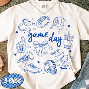 Op de afbeelding: Wit t-shirt met blauwe grafische illustraties van voetbalgerelateerde items, waaronder een voetbal, helm en megafoon. De woorden "game day" zijn in blauwe letters geschreven. Het shirt heeft ook een blauw-witte PNG-afbeelding.