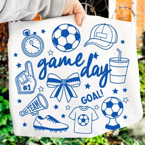 Może przedstawiać: Biała tkanina z niebieskimi ilustracjami o tematyce piłkarskiej, w tym piłka nożna, stoper, piankowy palec, megafon, kokarda oraz słowa "game day" i "goal!". Projekt zawiera r&oacute;wnież czapkę z daszkiem, kubek, but piłkarski i gwiazdy.