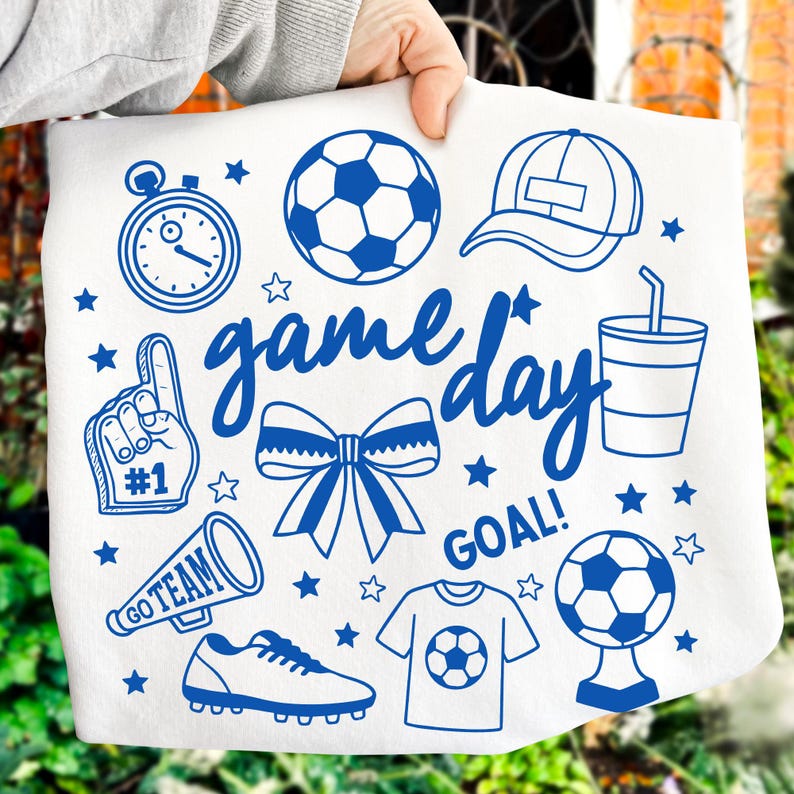 Może przedstawiać: Biała tkanina z niebieskimi ilustracjami o tematyce piłkarskiej, w tym piłka nożna, stoper, piankowy palec, megafon, kokarda oraz słowa "game day" i "goal!". Projekt zawiera r&oacute;wnież czapkę z daszkiem, kubek, but piłkarski i gwiazdy.