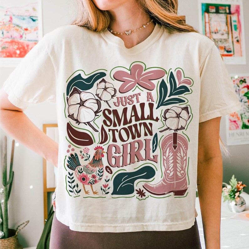 Pu&ograve; includere: T-shirt color crema con un design grafico. Il design presenta la scritta "JUST A SMALL TOWN GIRL", uno stivale da cowboy rosa, un gallo, batuffoli di cotone ed elementi floreali in rosa, verde e marrone.