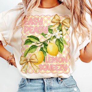 Pu&ograve; includere: Una maglietta bianca con un design grafico rosa e giallo con limoni, fiocchi e il testo "Easy Peasy Lemon Squeezy".