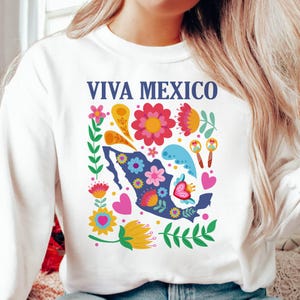 May include: White sweatshirt with a blue outline of Mexico and the text "Viva Mexico" in blue. The outline is surrounded by colorful flowers, hearts, and maracas.