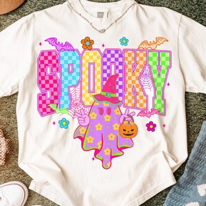 May include: A white t-shirt with a colorful "SP00KY" graphic. The design features a ghost wearing a witch hat and holding a jack-o'-lantern. Bats, flowers, and a spiderweb complete the Halloween theme.