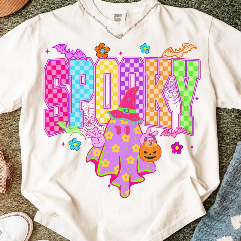 May include: A white t-shirt with a colorful "SP00KY" graphic. The design features a ghost wearing a witch hat and holding a jack-o'-lantern. Bats, flowers, and a spiderweb complete the Halloween theme.