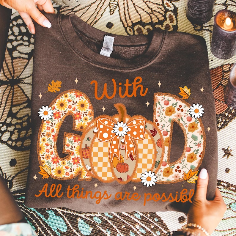 May include: A brown sweatshirt featuring the words "With God" and "All things are possible" in a fall-themed design. The design includes a pumpkin and floral-filled letters, with daisy accents. The sweatshirt is held by hands.