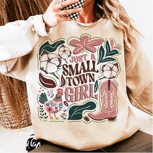 Pu&ograve; includere: Felpa beige con un design grafico con la scritta "Just a Small Town Girl". Il design include un gallo, fiori di cotone, uno stivale da cowboy ed elementi floreali nei toni del rosa, verde e marrone.