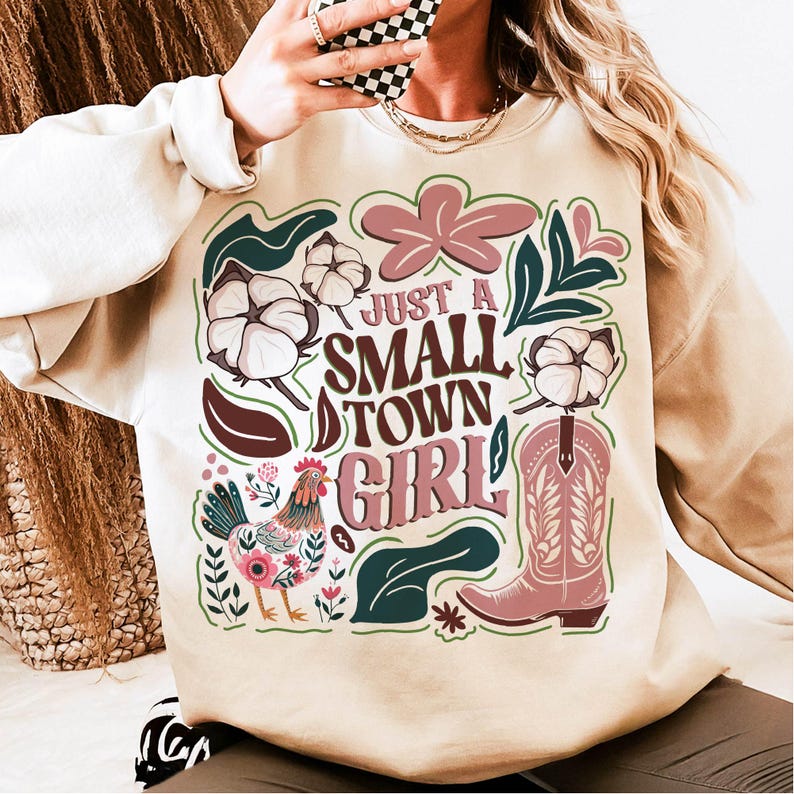 Pu&ograve; includere: Felpa beige con un design grafico con la scritta "Just a Small Town Girl". Il design include un gallo, fiori di cotone, uno stivale da cowboy ed elementi floreali nei toni del rosa, verde e marrone.