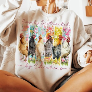 以下が含まれることがあります： クリーム色のスウェットシャツ。筆記体で「Easily Distracted by Chickens」の文字。カラフルな花のストライプの前に、6羽の水彩画の鶏が描かれています。鶏の色と模様は様々です。