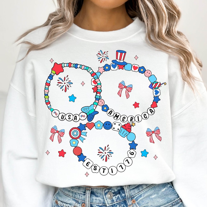 May include: White sweatshirt featuring a colorful graphic design with the words "USA AMERICA EST 1776". The design includes bracelets with stars, bows, and fireworks in red, white, and blue.