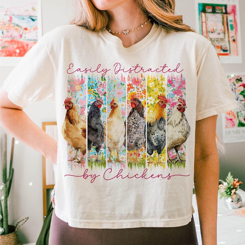 以下が含まれることがあります： クリーム色のTシャツ。6羽の鶏が花柄の背景の前に描かれた水彩画のデザイン。テキスト「Easily Distracted by Chickens」は、デザインの上部と下部に筆記体で書かれています。