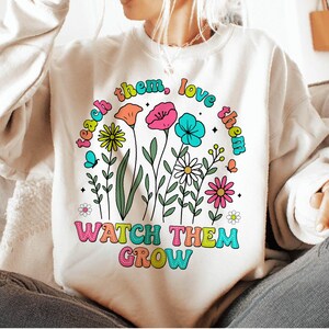 Puede incluir: Una sudadera blanca con un dise&ntilde;o gr&aacute;fico colorido que presenta flores y el texto "teach them, love them, watch them grow".