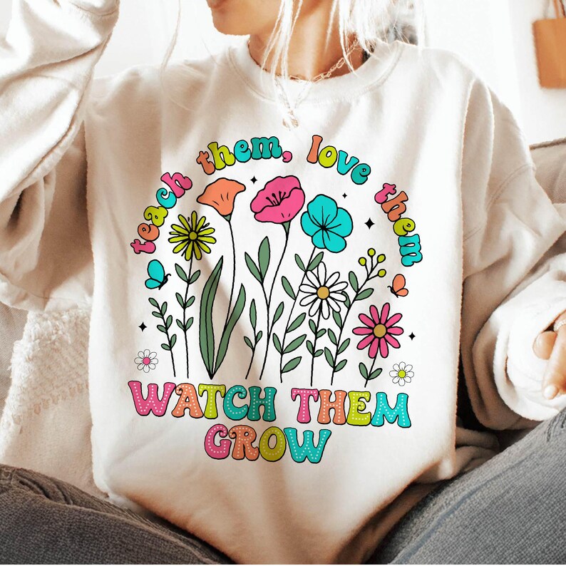 Puede incluir: Una sudadera blanca con un dise&ntilde;o gr&aacute;fico colorido que presenta flores y el texto "teach them, love them, watch them grow".