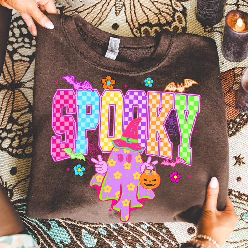 May include: A brown sweatshirt with the word "SPOOKY" in colorful checkered letters. The design includes a ghost character wearing a witch hat and holding a jack-o'-lantern, with bats and flowers. A fun Halloween-themed garment.