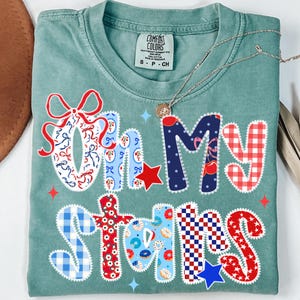 May include: A teal Comfort Colors t-shirt with the phrase "Oh My Stars" in a patchwork design. The letters are decorated with bows, stars, and patterns in red, white, and blue. A silver necklace with a small pendant rests on the shirt.